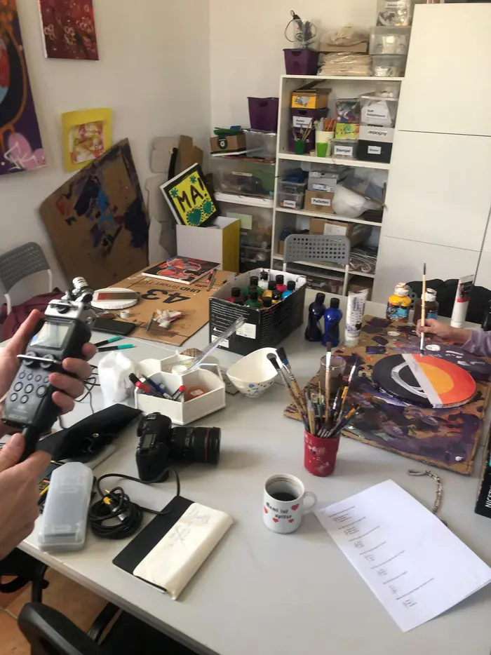 Ein vollgestellter Kreativarbeitsplatz mit Farben, Pinseln, Kameraequipment und einem Aufnahmegerät. Eine Person bemalt ein rundes Holzbrett.
