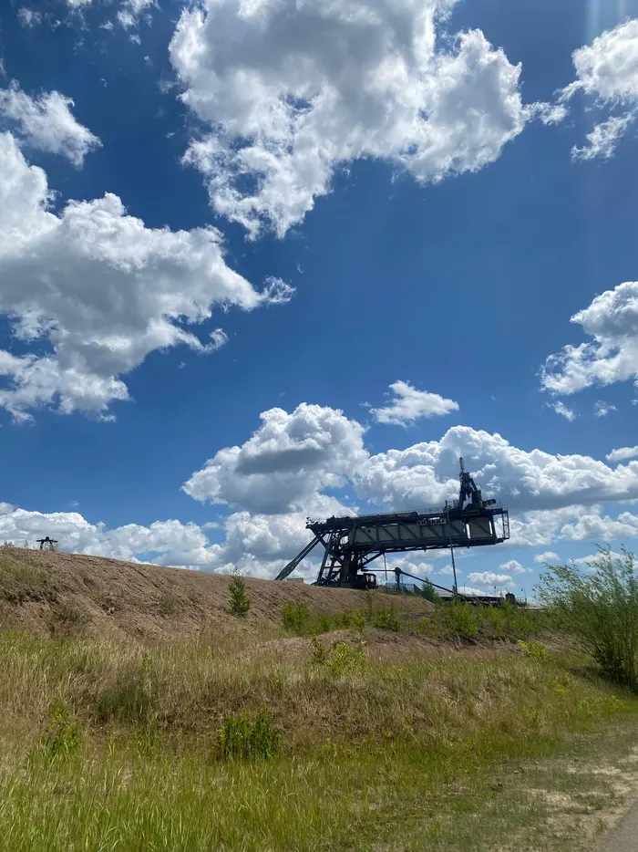 Eine weite, grüne Landschaft mit einer massiven, dunklen Förderbrücke im Hintergrund unter einem dramatisch bewölkten Himmel.