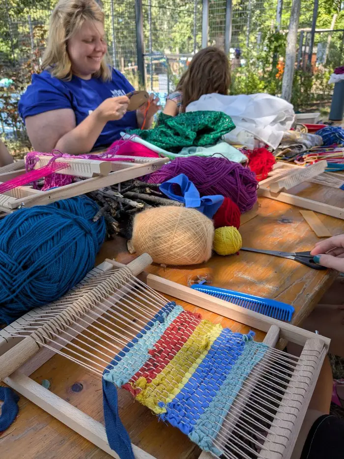 Eine lächelnde frau sitzen an einem Tisch mit vielen bunten Wollknäulen und Webrahmen. 
