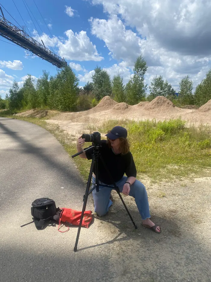 Eine Person kniet auf einem Weg und richtet eine Kamera auf einem Stativ aus. Im Hintergrund sind Sandhügel, Bäume und ein Teil einer alten Industrieanlage zu sehen.