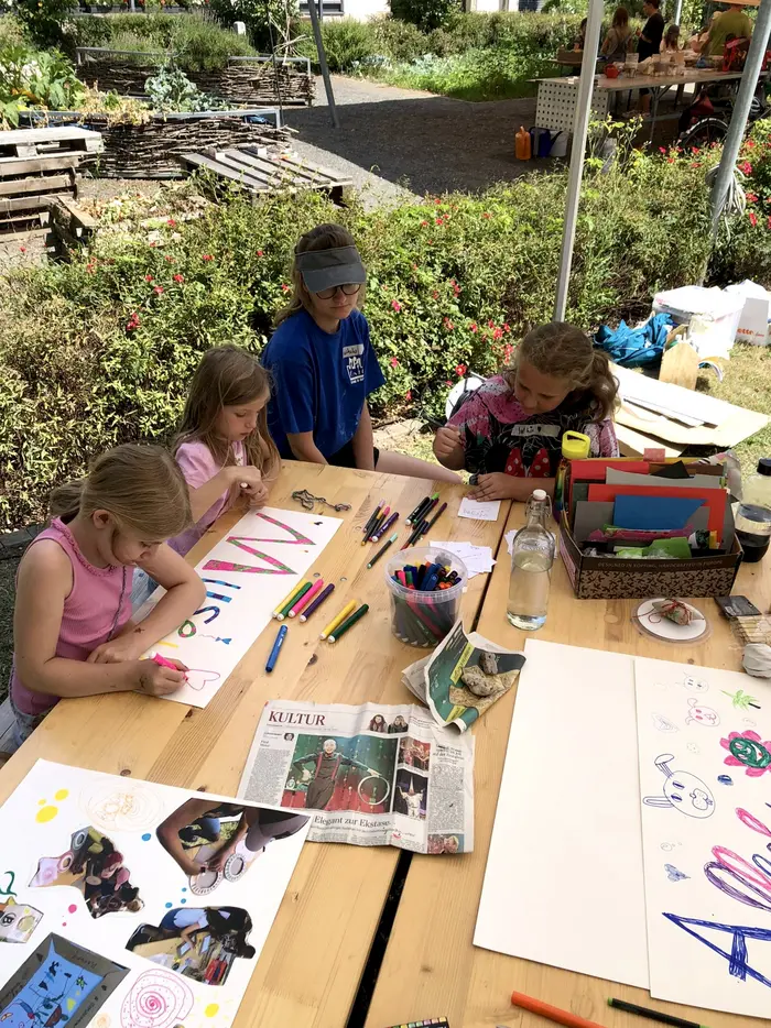 Drei Kinder und eine Frau sitzen an einem Tisch bei sommerlichen Wetter. Die Kinder zeichnen gemeinsam mit bunten Farben. Auf dem Tisch liegen Stifte und Papier.