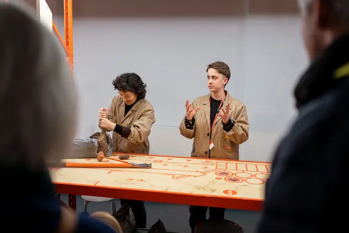 Zwei Personen in beigefarbenen Arbeitskitteln stehen hinter einem Tisch und führen ein Live-Format im Museum durch. Eine Person spricht gestikulierend, die andere arbeitet mit Ton. Im Vordergrund sind Besucher:innen zu sehen.