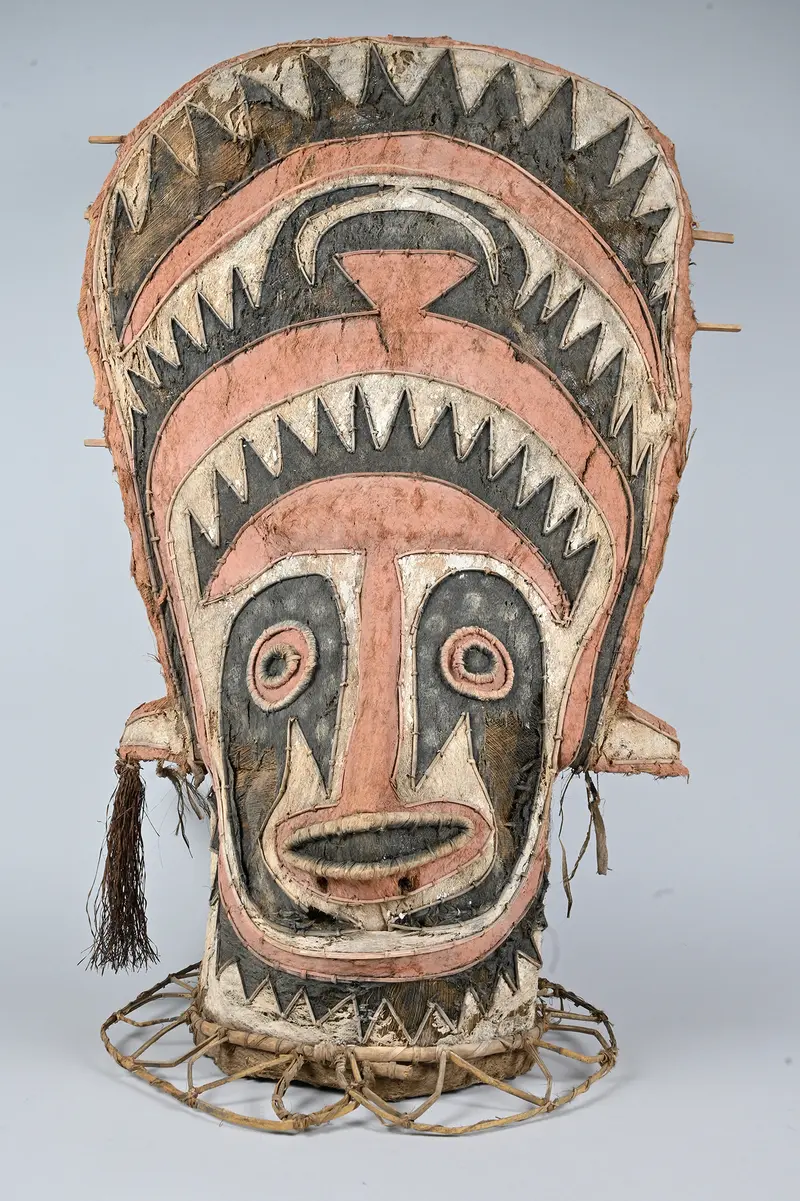 Eine große, dekorative Maske aus natürlichen Materialien, möglicherweise Leder oder Rindentuch, mit einem komplexen Muster und einem runden Rahmen an der Basis.