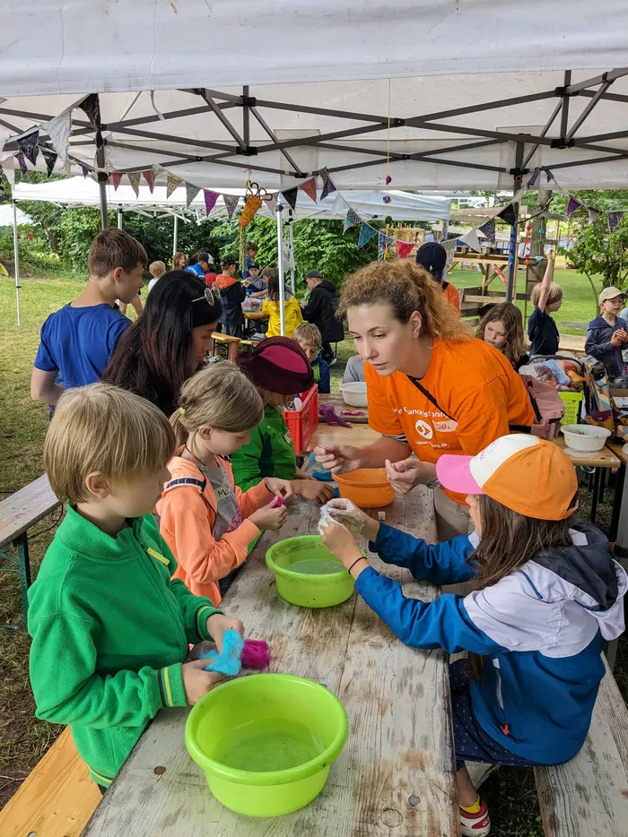 Kinder sitzen unter einem Pavillon an Tischen und arbeiten gemeinsam mit Betreuenden an einem Filzprojekt. Es herrscht eine kreative und fröhliche Atmosphäre bei einem Mitmachangebot im Freien.