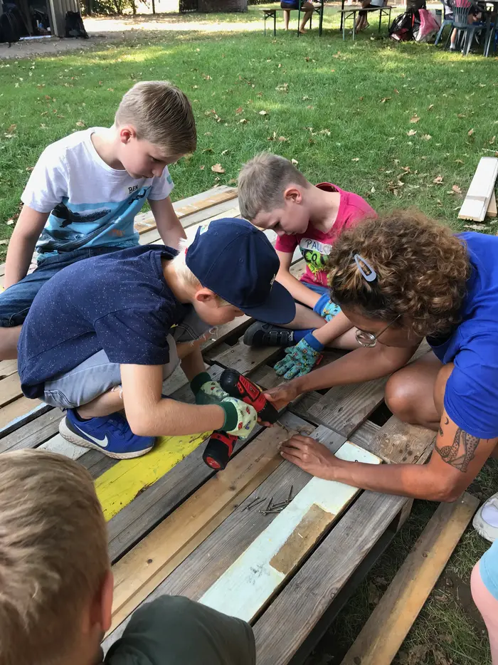 Mehrere Kinder und eine erwachsene Betreuungsperson schrauben gemeinsam Holzbretter auf einer Wiese zusammen. Die Kinder arbeiten konzentriert mit Akkuschraubern im Rahmen eines Bauprojekts.